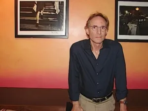 Sandy Kreisberg, aka HBSGuru, as he talks about Dee's interview, stands against a wall with two framed, professional black and white photographs are hung won either side of him.