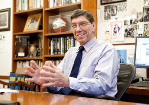 Photo of Harvard's Clay Christensen, sitting at a desk, gesturing with his hands while talking.