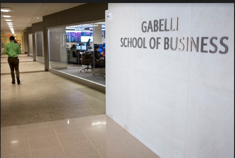 Interior hallway with Gabelli School of Business sign on wall to the right. Beyond sign there is a wall made of windows that has computers on the other side. The back of a man walking down the hallway can be seen in the background.