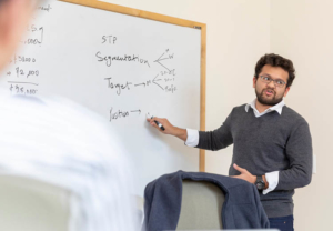 Man with dark hair, beard, glasses in front of whiteboard teaching a Gies business class.