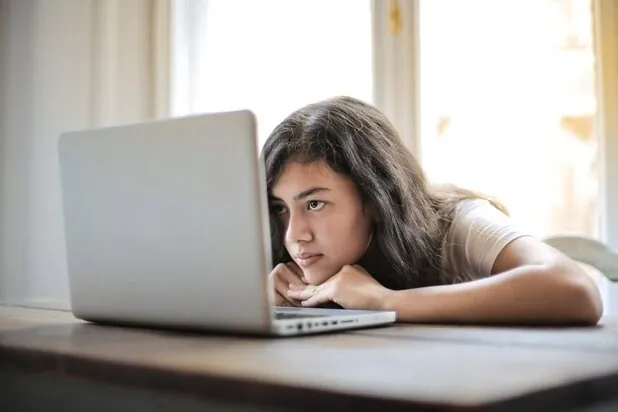 girl resting chin on table looking at laptop with a bored expression on her face